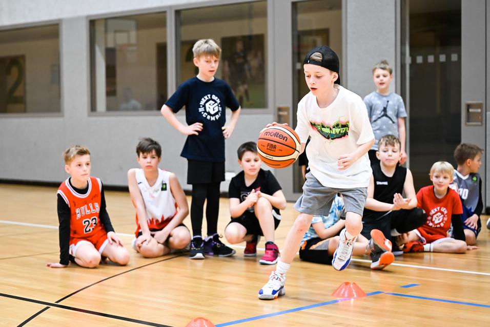 Ein Junge dribbelt einen Basketball auf einem Sportboden, während andere Kinder im Hintergrund zuschauen.