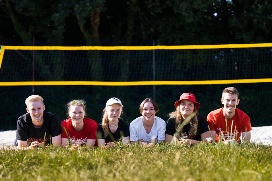 Sechs Personen liegen im Gras vor einem Beachvolleyballfeld mit gelben Netzen und Bäumen im Hintergrund.