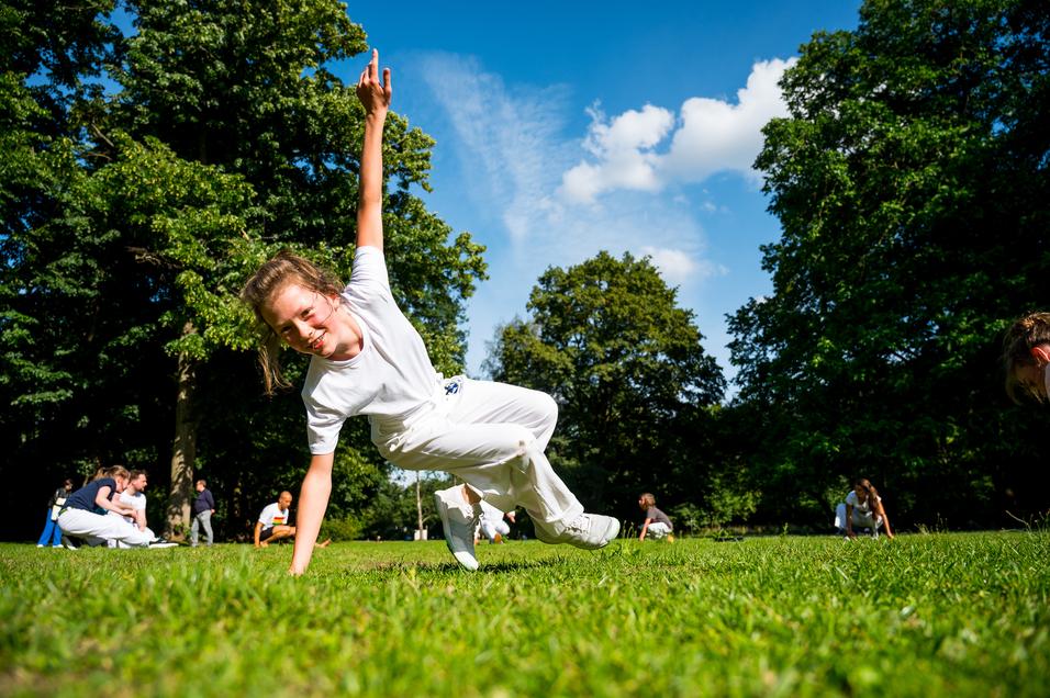 Mädchen in weißem Outfit zeigt eine Sportübung auf dem Rasen, umgeben von anderen Personen im Hintergrund.