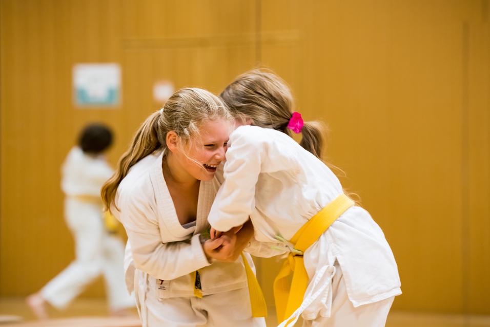 Zwei Mädchen im Judoanzug lachen fröhlich miteinander auf einer Judomatte. Eines trägt einen gelben Gürtel.