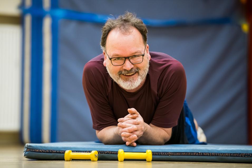 Mann mit Brille und Bart liegt auf einer Matte, lächelt und hat zwei gelbe Hanteln vor sich.