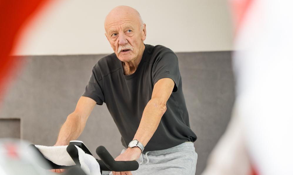 Älterer Mann auf einem Spinning-Bike, konzentriert, mit Hand auf dem Lenker und Handtuch auf dem Sattel.