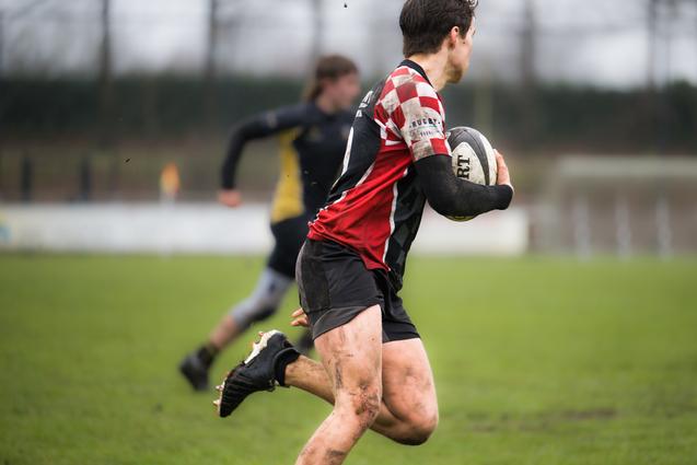 Spieler in gestreiftem Rugbytrikot rennt mit einem Rugbyball über ein grasbedecktes Spielfeld, leicht verschlammt.