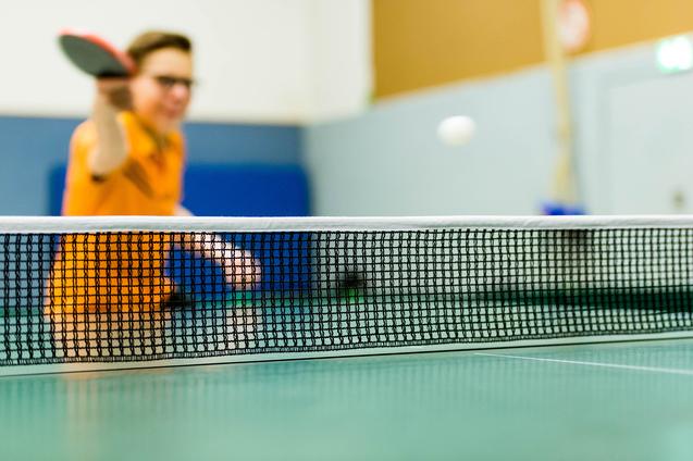 Tischtennisschläger in der Hand eines Spielers, im Hintergrund ein Ball über das Netz fliegend auf einer grünen Tischtennisplatte.